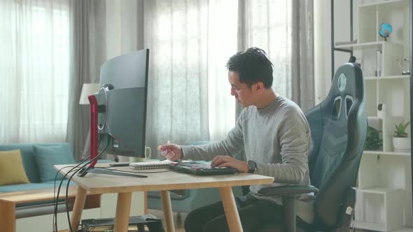 Asian Man Looking At Computer And Writing In Notebook While Working At Home alt