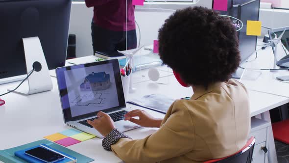 Mixed race businesswoman wearing face mask sitting using a laptop in modern office alt