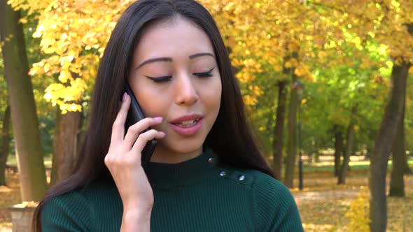 A Young Asian Woman Talks on A Smartphone in A Park alt