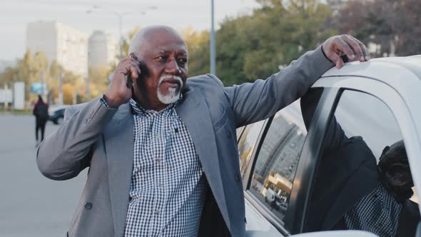 Old African American Man Stands Near Automobile Communicates on Telephone in Street Parking Simple alt