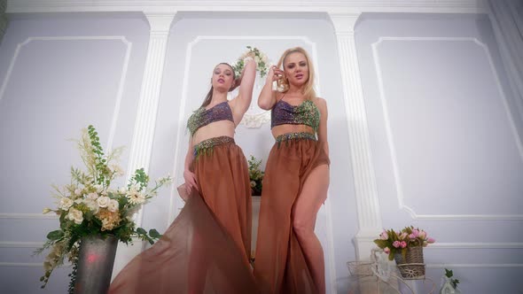 Two Women in Evening Dresses Dance and Sing in a Large Blue Room with Flowers