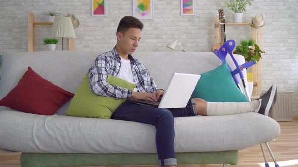 Asian Young Man with Broken Leg in Bandages Lying on Sofa with Laptop alt