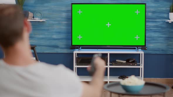 Over Shoulder View of Man Switching Channels While Looking at Green Screen on Tv and Sitting on Sofa alt