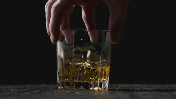 Hand Holds Glass with Golden Whisky. Whiskey with Ice Cubes Rotating in Glass. alt