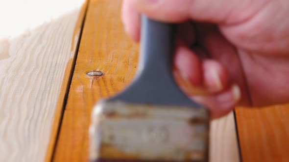 A man paints freshly planed wooden board with a brush on the window background on the workshop