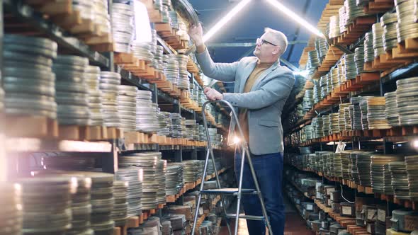 Archive Worker is Standing on a Ladder and Choosing a Film Tape alt