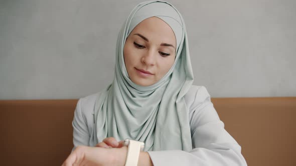 Cheerful Modern Muslimah Looking at Smart Watch Screen and Smiling Indoors in Cafe alt