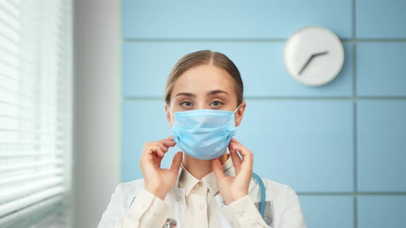 Blonde young woman nurse in white coat puts on blue disposable facial mask alt