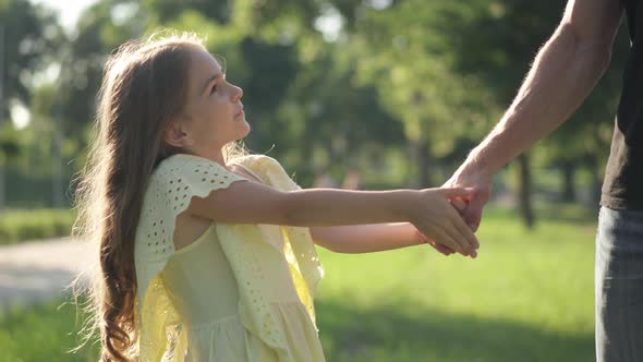 Charming Brunette Little Girl in Yellow Dress Pulling Male Hand Talking Standing in Sunbeam Outdoors alt