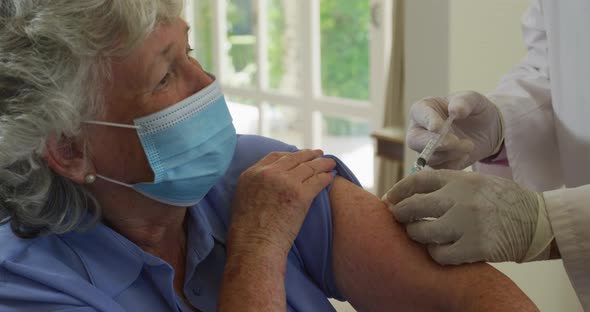 Caucasian senior male doctor wearing face mask injecting covid-19 vaccine into senior woman at home alt