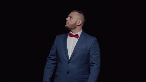 An elegant man in a blue tuxedo with a red tie on a black background, looks up and then forward alt