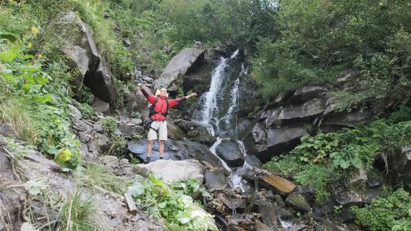 Tourist with Backpack Came To Mountain Waterfall and Spreading Arms To Sides alt