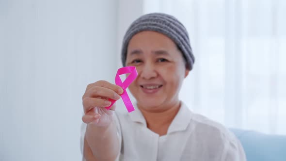 Elderly female patient holding pink ribbon in hospital. Breast cancer awareness concept. alt