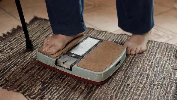 Senior sick woman stands on an old dirty scale and measures her weight. alt