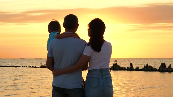 A Happy Family Enjoys the Sunset Over the Sea. A Father Holds His Son in His Arms. Mother and Son alt