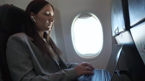 Brunette Woman in a Suit and a Wireless Earphone in Ear is Typing Text on a Laptop While Sitting on alt
