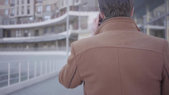 Back View of Handsome Successful Man in Brown Coat Walking on the City Street Talking By Mobile alt