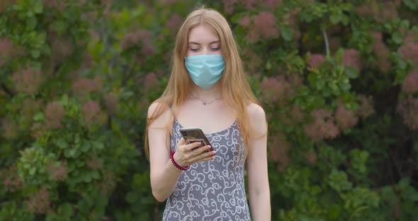 A young girl in a protective medical mask looks at the phone and sends messages