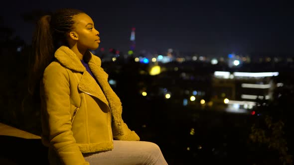 A Young Black Woman Looks Around and Smiles at the Camera As She Sits in an Urban Area at Night alt
