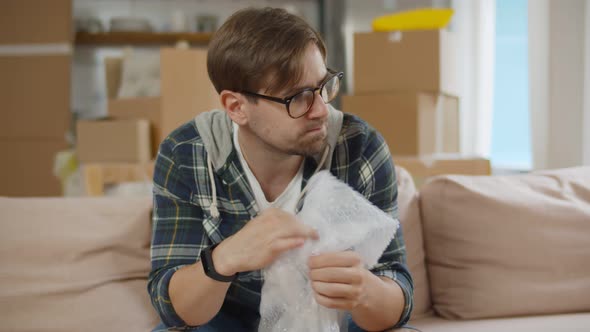 Stressed Young Man Popping Bubbles of Bubble Wrap Worried About Moving in New House alt