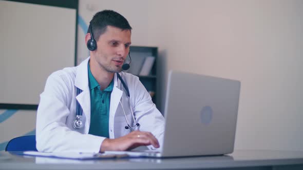 Friendly Male Doctor in White Medical Coat With Headphones Making Conference Call on Laptop alt