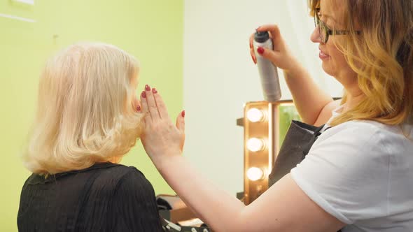 Professional Hairdresser, Stylist Doing Hairstyle for Senior Woman Spraying with Lacquer for Fixing alt