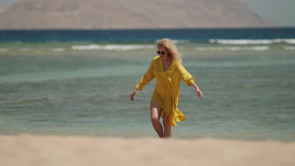 a Curlyhaired Blonde in Sunglasses and a Yellow Fluttering Dress Waves Hands Takes Sandals and Goes alt