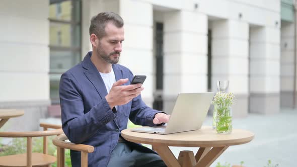Man Browsing Internet on Smartphone While Working on Laptop alt