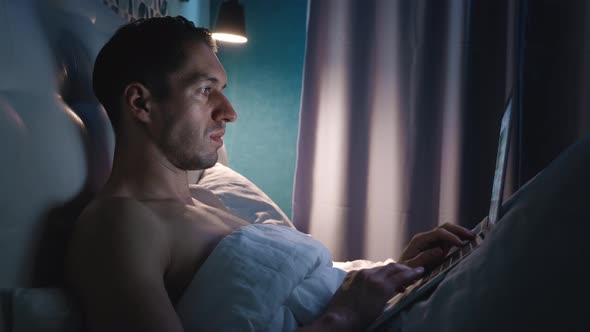 Portrait of Man Lying on a Bed in the Bedroom at Nighttime at Home and Using Laptop alt