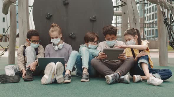 Preteens in Face Masks Using Gadgets at Playground alt