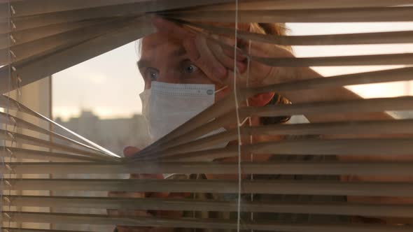 Worried Man in Protective Mask Looking through Window Blinds during Flu Quarantine
