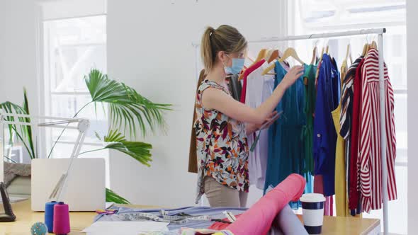Female fashion designer wearing face mask looking checking on dresses at studio alt