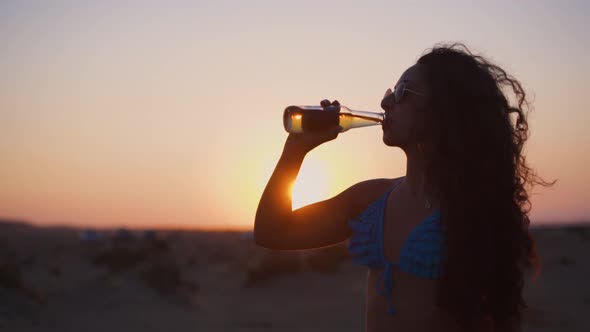 Hipster Girl in Bikini and Sunglasses Drinking Alcohol at Sunset