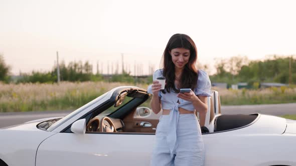 Beautiful Stylish Woman Leans on Her Luxury White Convertible and Keeps Cup of Coffee