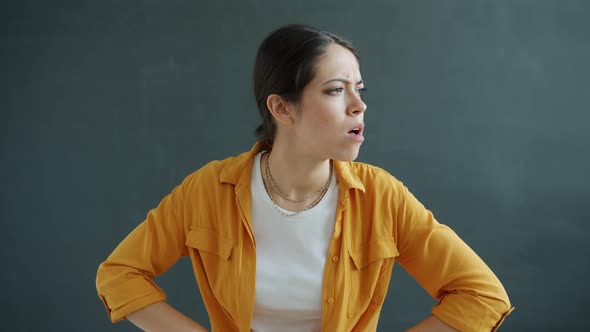 Portrait of Unhappy and Angry Young Woman Looking at Camera Standing Akimbo on Gray Background alt