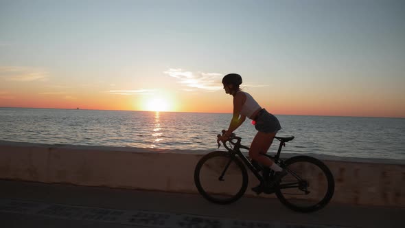 Young female is cycling on bicycle along sea promenade at sunrise. Woman cycling outdoors alt
