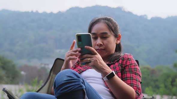 Pretty Asian woman sits on a camping chair and uses a smartphone to communicate during