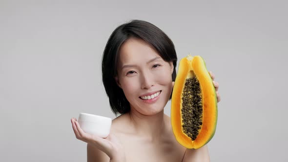 Mature Japanese Woman Holding Papaya And Moisturizer Over Gray Background alt