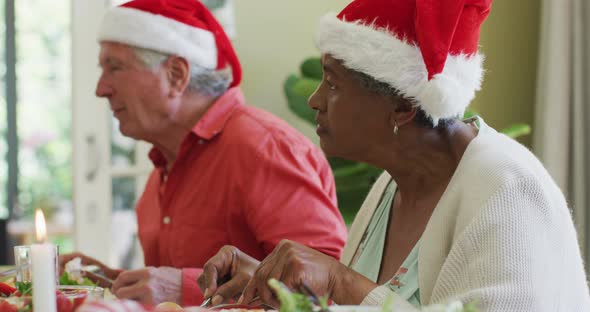 Diverse senior man and woman in santa hats listening and eating at christmas dinner table at home alt