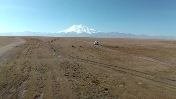 Tourist White Car Fast Driving on Off Road Way on Highland Field on Snowy Elbrus Mount Landscape alt