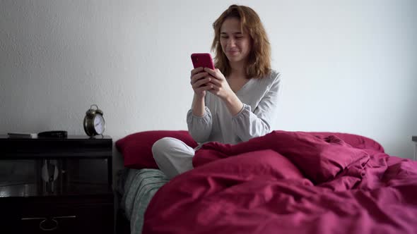 Young Woman Sitting in Bed in Pajamas Interacting with Mobile Phone
