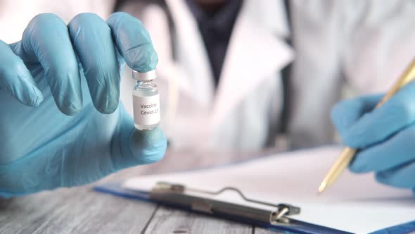 Hand in Latex Gloves Holding Coronavirus Vaccine and Writing on Paper alt