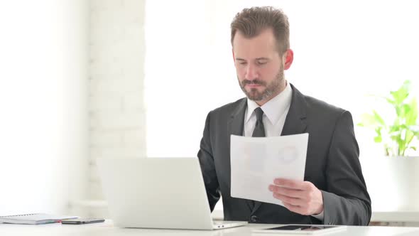 Young Businessman with Laptop Reading Documents in Office alt