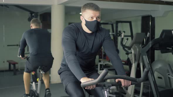 A Young Man in Sportswear Exercises a Stationary Training Bike in a Gym alt