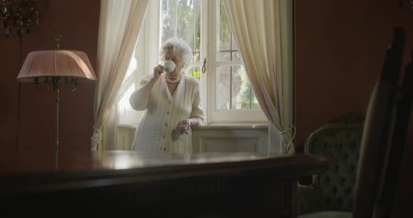 Senior Grandma Woman Enjoying Drinking Cup of Tea or Coffee Near Window