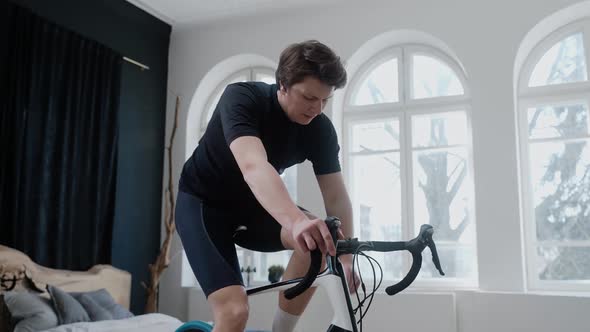 Side View of Cyclist Training on an Exercise Bike and Drinking Water at Home alt