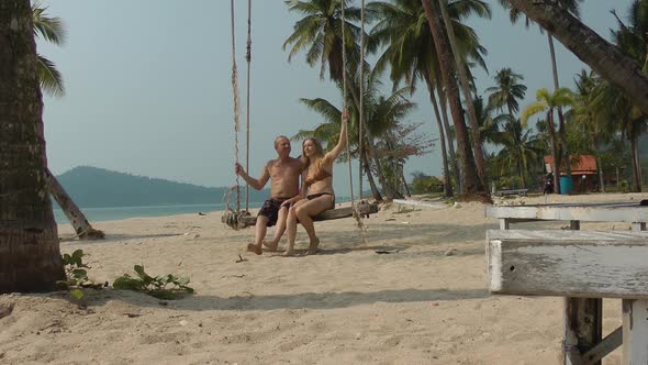 a Man and a Woman in a Swimsuit Swing on a Swing Suspended From a Palm Tree on the Beach of the Sea
