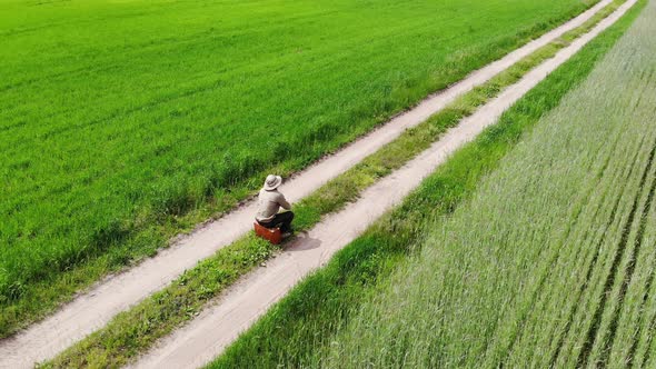 Man Alone Walking By Country Road Make a Stop in a Green Field for a Rest and Sit on His Suitcase