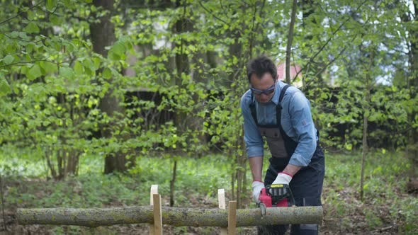 Lumberjack in Workwear Saws Firewood on Sawhorses with Electric Saw. Forest. alt