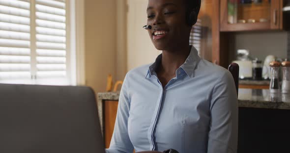 African american woman wearing phone headset having a videocall on laptop while working from home alt